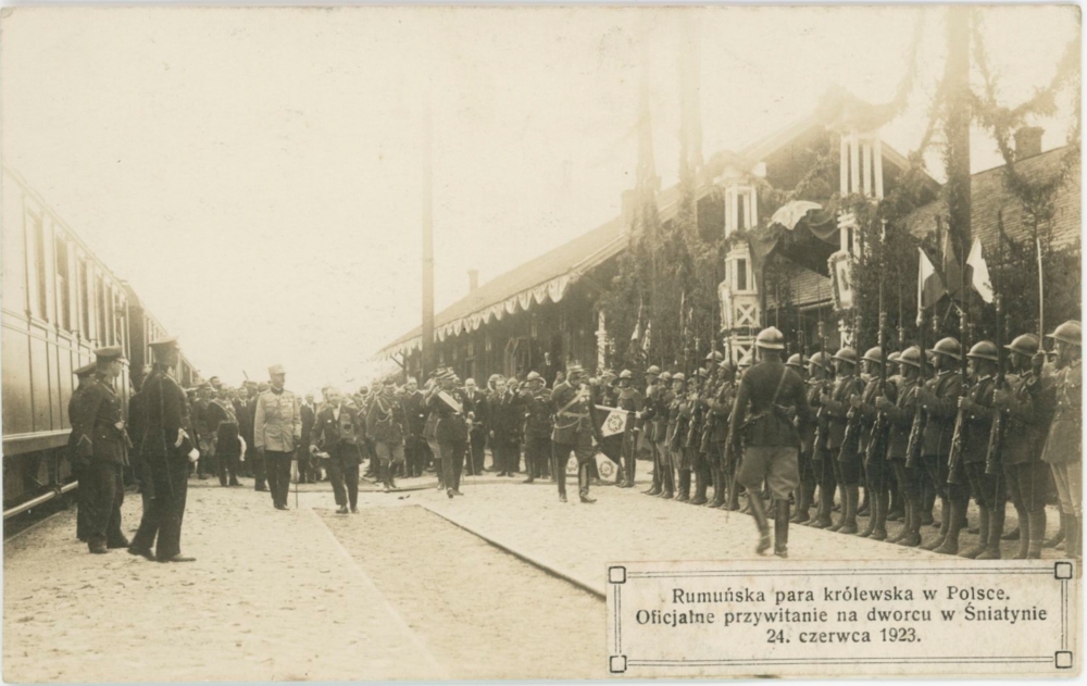 Zdjęcie prezentuje fragment budynku dworca, a lewej strony część składu pociągu. W centralnej części fotografii znajdują się żołnierze polscy i rumuńscy oficerowie, a wśród nich król Ferdynand w szarym mundurze, czapką na głowie, z podniesioną prawą ręką.&nbsp;