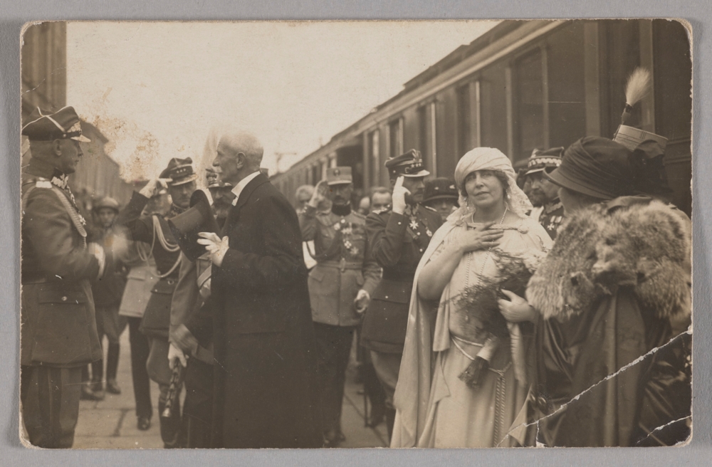 Fotografia przedstawia powitanie pary królewskiej na dworcu w Warszawie. w centralnej części widoczna jest królowa Maria w jasnym stroju. Tyłem w kapeluszu na głowie stoi prezydentowa Maria Wojciechowska. na drugim planie znajduje się prezydent Stanisław Wojciechowski trzymający cylinder w dłoni tuż za nim stoi król Ferdynand I. Wokół salutują oficerowie.