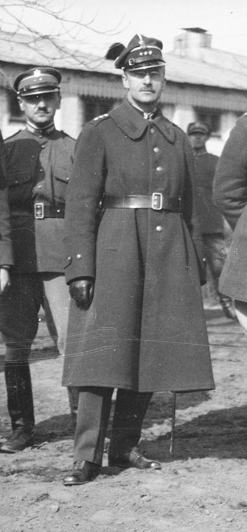 Na zdjęciu prezentuje się mężczyzna w wojskowej kurtce i czapce. Na czapce widnieje orzełek, a pod nim trzy gwiazdki. Za nim, w tle widoczni są inni żołnierze.
