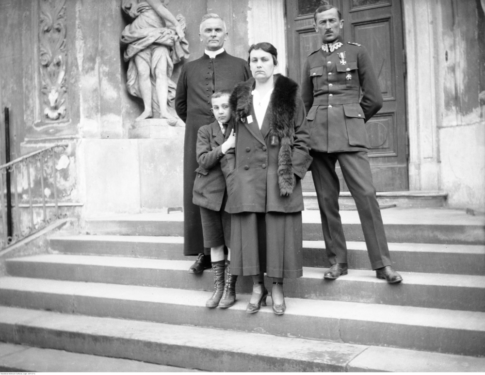 Fotografia przedstawia ks. Mariana Tokarzewskiego ubranego w sutannę. Ksiądz Marian Tokarzewskie wraz z niezidentyfikowanymi osobami stoi na schodach przed budynkiem.