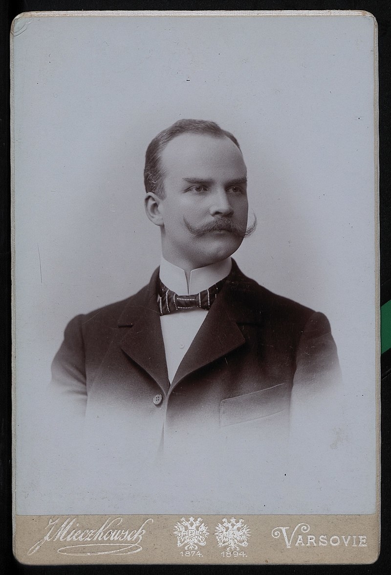 Fotografia Emila Młynarskiego prezentującego się w marynarce. Mężczyzna krótkie zaczesane na bok włosy i długie zakręcone ku górze wąsy.