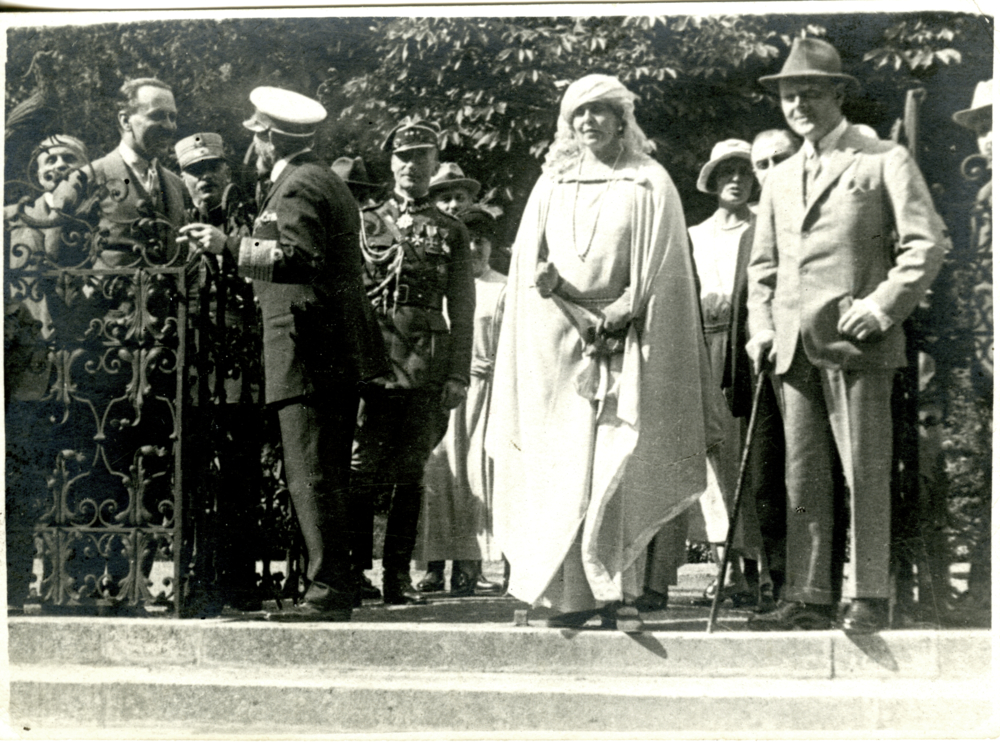 Na fotografii widoczne są osoby elegancko ubrane. Król w mundurze admirała stoi odwrócony. Obok niego stoi królowa ubrana w biały strój. Alfred Potocki stojący z prawej jest ubrany w garnitur oraz kapelusz.